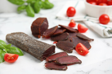 Delicious dry-cured beef basturma with basil and tomatoes on white marble table, closeup
