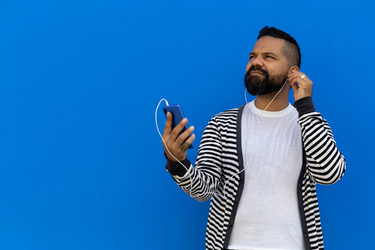 Adult Male (39) With Beard, Medium Flat, Listens To Music From His Cell Phone Holding A White Earphone To His Ear. He Looks Up From His Right Side. Dressed In Striped Sweater, White T-shirt, Blue Wall