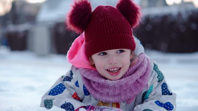 Close up Portrait Little Child Pretty Girl Kid Smiling Looking at Camera play red hat lie on Stomack. Snow Park Outdoor. Winter time, happiness concept Snowing Cold frosty weather Carefree childhood