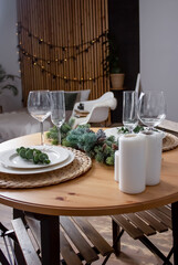 Round table decorated for Christmas and New Year. Bed in the background. Christmas tree twigs on the table. Plates and wine glasses on the table. Kitchen set and flowers on the background. 
