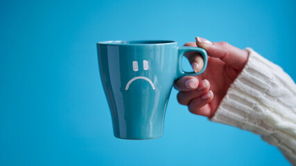 Blue Monday concept. Close-up of a female hand in a white sweater holds a blue cup with a sad...