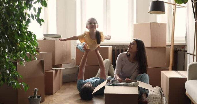 Happy young family rest on warm floor on relocation day in living room near heap of cardboard boxes, dad lifts on outstretched hands cute preschooler 6s daughter. New house, move, leisure, fun concept