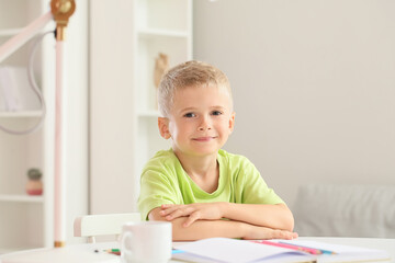 Naklejka premium Smiling little boy sitting at table with drawing paper and colored pencils in bright bedroom or playroom, cute child portrait at home with copy space