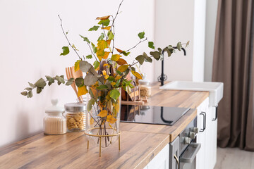 Vase with beautiful autumn branches and jars with food on wooden counter in light kitchen