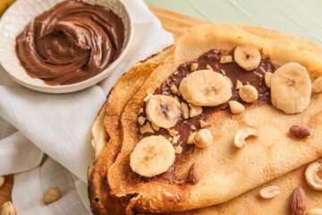 Board of tasty thin pancakes with chocolate paste, cut banana and nuts on table, closeup