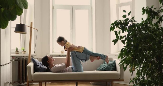 Mom lying on sofa raise on hands little daughter, girl spread arms like airplane wings, imagines like flying on air, enjoy playtime at home with mother. Family fun and weekend leisure, travel concept