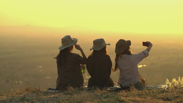 A group of Asian young women who are friends are having a good time and enjoying themselves. Sit together enjoy the sunset while taking a selfie on the mountain, outdoor camping trip. slow motion