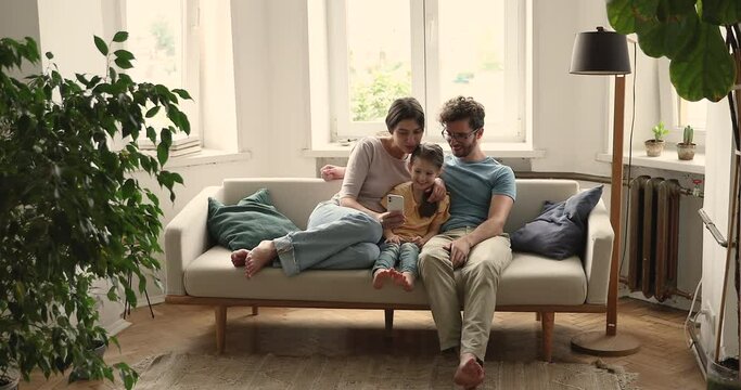 Couple and 5s daughter spend time together sit on sofa with cellphone use new mobile app, having fun enjoy internet amusing content enjoy carefree weekend. Family leisure at home, modern tech concept