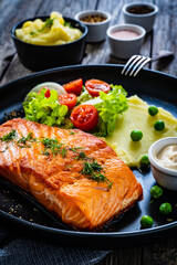 Fried salmon steak with potato puree and vegetable salad served on black plate on wooden table
