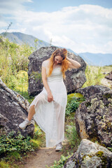 Young cheerful girl in white dress posing on the background of mountains on summer day. Brunette having fun in nature. Vertical photo.