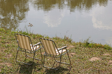 Fototapeta premium Pair of empty folding garden chairs on grass next to the edge of a river or a lake on a sunny summer day. No people.