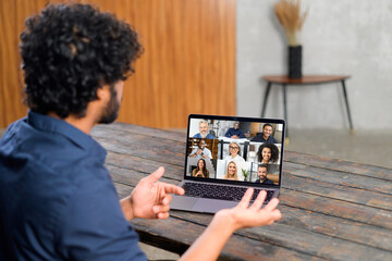 Remote meeting via video call. Indian man sitting in the kitchen and using app on laptop for online communication with group of diverse multiracial people. Back view