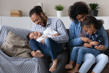 Excited joyful African parents cuddling, tickling kids on couch, giggling, laughing, enjoying...