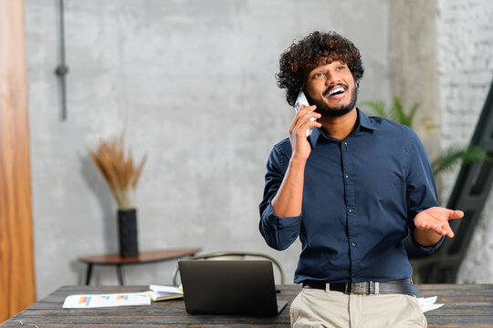 Happy middle eastern man talking on the smartphone while standing in modern loft office. Smiling successful contemporary man manager in casual wear holding phone call