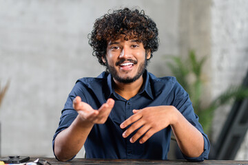 Portrait of happy young Indian guy sitting at desk and gesturing. Smiling curly ethnic man in...