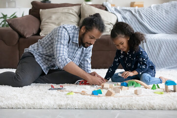 Sweet African preschool kid girl and focused dad constructing city road model, playing with toy wooden blocks, cubes on soft carpeted floor in home living room, improving creative skills. Fatherhood