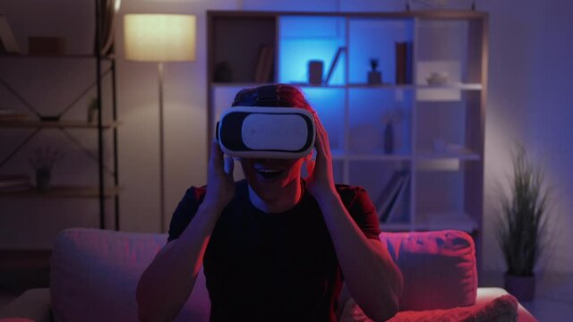 Male gamer. Virtual reality. Cyber entertainment. Surprised man in vr goggles feeling amazed and happy sitting sofa dark neon light home interior.