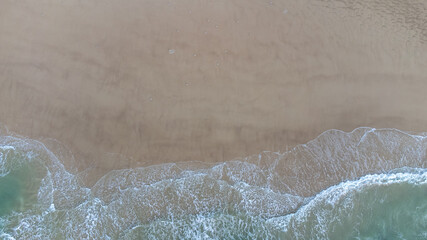 Aerial drone view perpendicular to the blue waves crashing on the sandy beach at the coast. The impact with wet wax creates white foam. The winter beach is empty. High quality photo