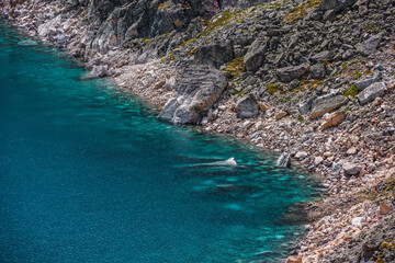 Top view to azure mountain lake with shiny cyan water edge among sunlit rocks. Beautiful alpine lake with stony bottom in transparent water in bright sun. Nature background of blue lake in sunlight.
