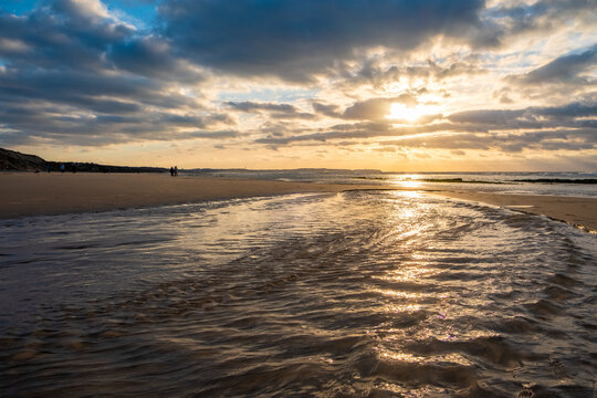 Amazing colorful sunset on a sandy beach at the sea under a sky painted with clouds and a golden sun. Picturesque nature scenery.Clouds reflected in water. Zen-like tranquil atmosphere no people