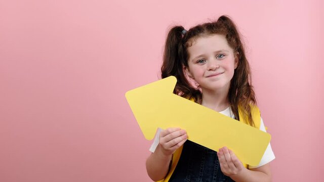 Smiling cute schoolgirl kid showing aside with yellow arrow, wears backpack, posing isolated over pink color background wall in studio with copy space for promotional content. Back to school concept