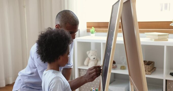 Happy Black father involve small son kid in education activity teach to make drawings on blackboard using chalks. Young male teacher babysitter give private lesson to little boy pupil in form of game