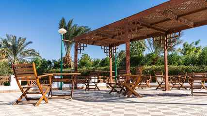 In the recreation area, under a lattice canopy, there are wooden chairs and tables. Shadows and sun on the tiled floor.  There is green vegetation all around. Palm trees against the blue sky. Egypt