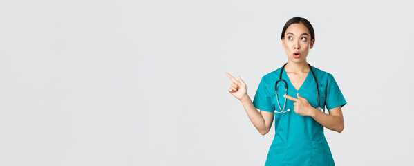 Covid-19, healthcare workers, pandemic concept. Intrigued and amazed beautiful asian female intern, nurse in scrubs or surgeon pointing and looking upper left corner, saying wow in amazement