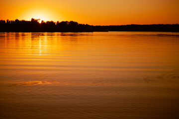 Fototapeta premium Shining lake with sun setting below horizon and covering with rays water surface with dark trees in background. Astonishing view full of yellow and orange colors.