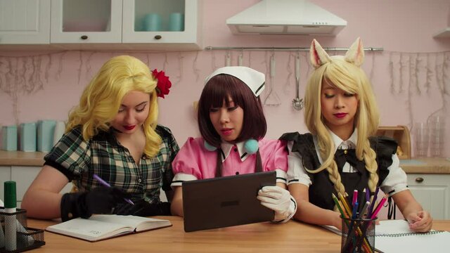Charming cheerful diverse multiracial female cosplayers wearing anime and comic book character costumes, networking online on digital tablet, talking and discussing cosplay performance at home.
