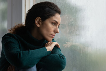 Thoughtful stressed young hispanic latin woman sitting on windowsill, looking outside on rainy...