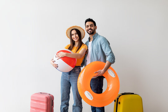 Happy couple ready for summer vacation, posing with inflatable circle and ball in hands over light wall