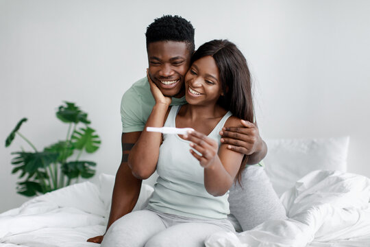 Happy young pretty african american wife and husband on bed in bedroom look at pregnancy test and enjoy good news - Powered by Adobe