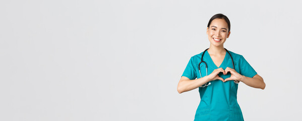 Covid-19, healthcare workers, pandemic concept. Lovely caring asian doctor, female nurse in scrubs showing heart gesture and smiling, taking care of patients with love, white background - obrazy, fototapety, plakaty