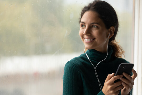 Dreamy peaceful happy young woman listening favorite music or affirmations in wired earphones from mobile playlist, standing near big window, copy space, hobby relaxation activity, inspiration concept