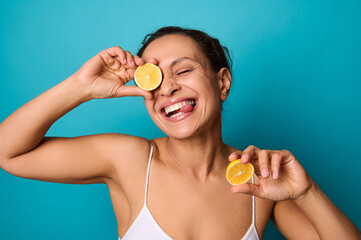 Attractive delighted young woman smiling with cheerful beautiful healthy toothy smile, holding lemon halves and covering her eyes with it. Isolated on bright blue background with copy ad space