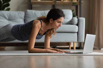 Fototapeta na wymiar Concentrated young beautiful sporty woman in activewear standing in plank on elbows, watching online class, working out or practicing yoga distantly, involved in web camera training at home.