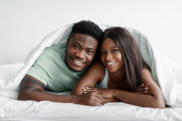 Fototapeta na wymiar Happy young attractive african american guy and lady lie on bed under covers in bedroom