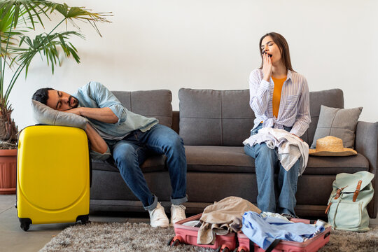 Young couple preparing for vacation, woman packing suitcases and yawning while husband sleeping on sofa