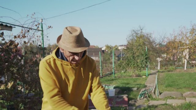 Senior farmer in brown hat sweeps autumn leaves with broom in hands at backyard of countryside house