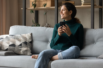 Relaxed young happy latin hispanic woman sitting on cozy sofa, looking in distance thinking of...
