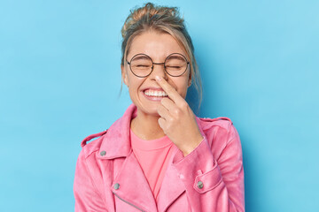 Cheerful woman with combed hair touches nose foolishes around wears spectacles and pink jacket isolated over blue background tries to amuse someone. Positive fair haired girl has fun indoor. - obrazy, fototapety, plakaty