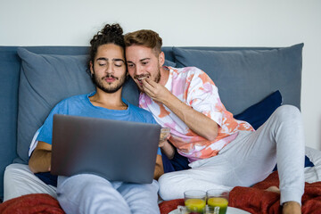 Young couple of same-sex people having breakfast in bed, gay couple of men watching a movie on laptop, video call in lockdown and quarantine due to Covid19 epidemic