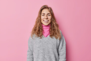 Fototapeta premium Beautiful happy woman with long hair smiles gladfully has clean natural skin ni make up feels happy wears casual jumper poses against pink background. Positive human face expressions and feelings