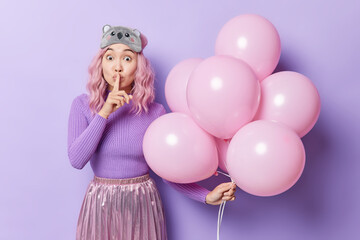 Surprised mysterious young Asian woman with dyed hair tells secret makes hush gesture holds bunch of inflated balloons wears casual clothes sleepmask isolated over purple background. Partying concept