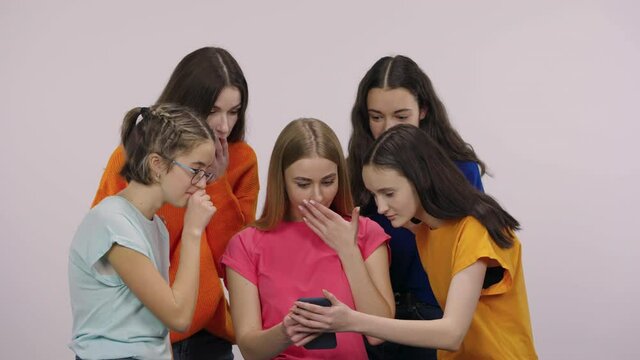 A group of pretty young girls looking at interesting news on their phones with wow delight and surprise. Five girls models posing on a white background in the studio. Close up. Slow motion.