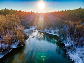 Drone view of a river with a lake on a snowy forest plain during a clear winter morning and bright sunrise