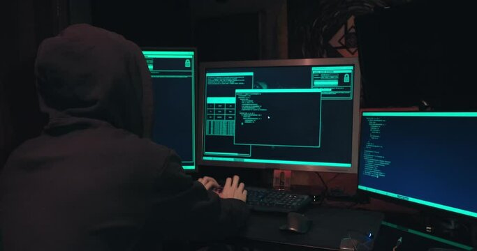 Hacker in a hood sits in front of computer screens and hacks databases. The concept of cyberterrorism, server hacking and cyberattacks. The room is dark, a person is typing code into the command line