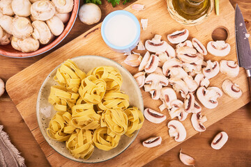 Ingredients for cooking traditional italian pasta with mushroom sauce - sliced champignons on a kitchen board, fettuccine and cream in a jug, top view, flat lay