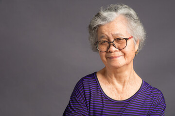 An elderly Asian woman looking at the camera with a smile while standing over a gray background